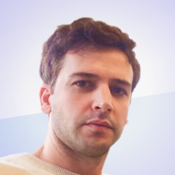 A man with short brown hair and a light beard is pictured against a gradient background that transitions from blue to light purple. He is wearing a light-colored top and looking directly at the camera with a neutral expression.