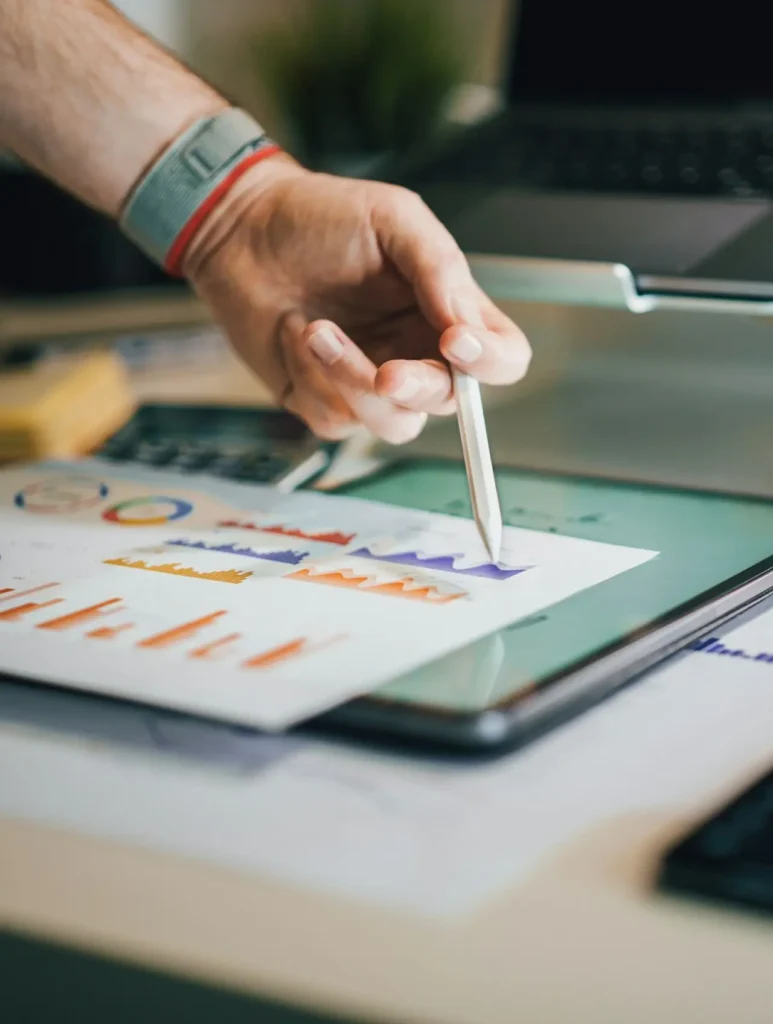 A person wearing a smartwatch points with a pen at a printed chart of colorful graphs and data, showcasing haro link building strategies. The paper rests on a laptop screen, suggesting data analysis or a business presentation.