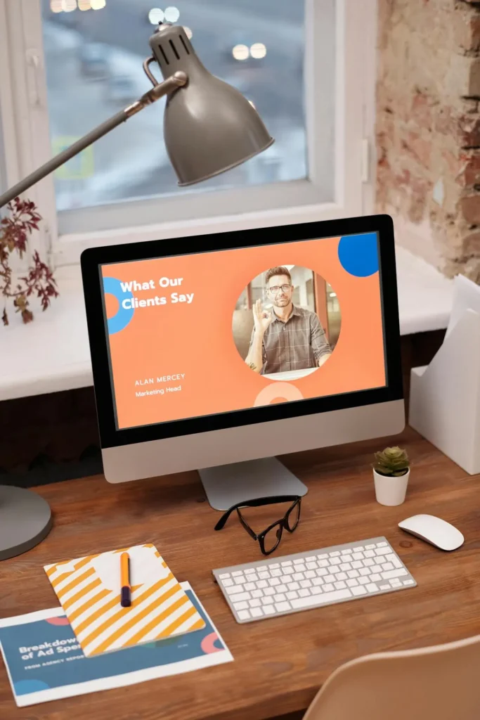 A workspace featuring a computer with a presentation slide titled "What Our Clients Say" and a person's photo. A lamp, glasses, plant, and documents clutter the wooden desk, hinting at strategic tasks like HARO link building. A window and brick wall create an inspiring background.