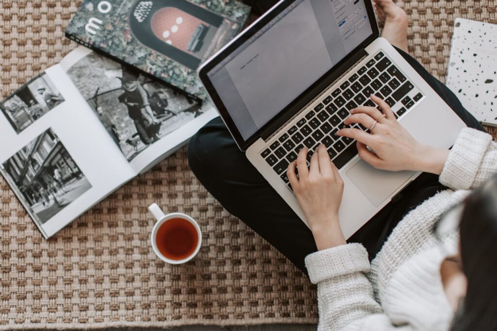 A person sits on the floor, immersed in crafting a HARO link building strategy. Surrounding them are open books filled with black-and-white photos and a steaming cup of tea on a patterned carpet. They wear a cozy sweater as they type diligently on their laptop.
