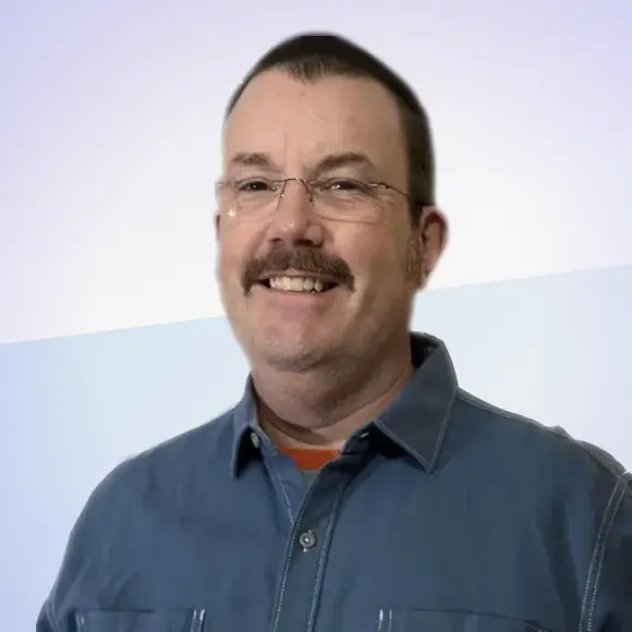 A man with glasses and a mustache smiles at the camera. He is wearing a blue button-up shirt. The background is a simple gradient of light tones.