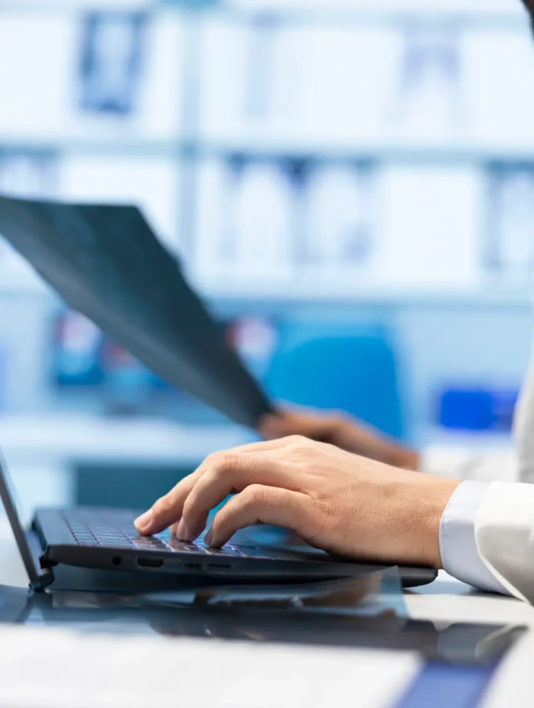 A close-up of a person's hands typing on a laptop keyboard, with a blurred background showing shelves and a blue chair. One hand is holding a sheet of paper, suggesting multitasking or reviewing documents while working on a computer.