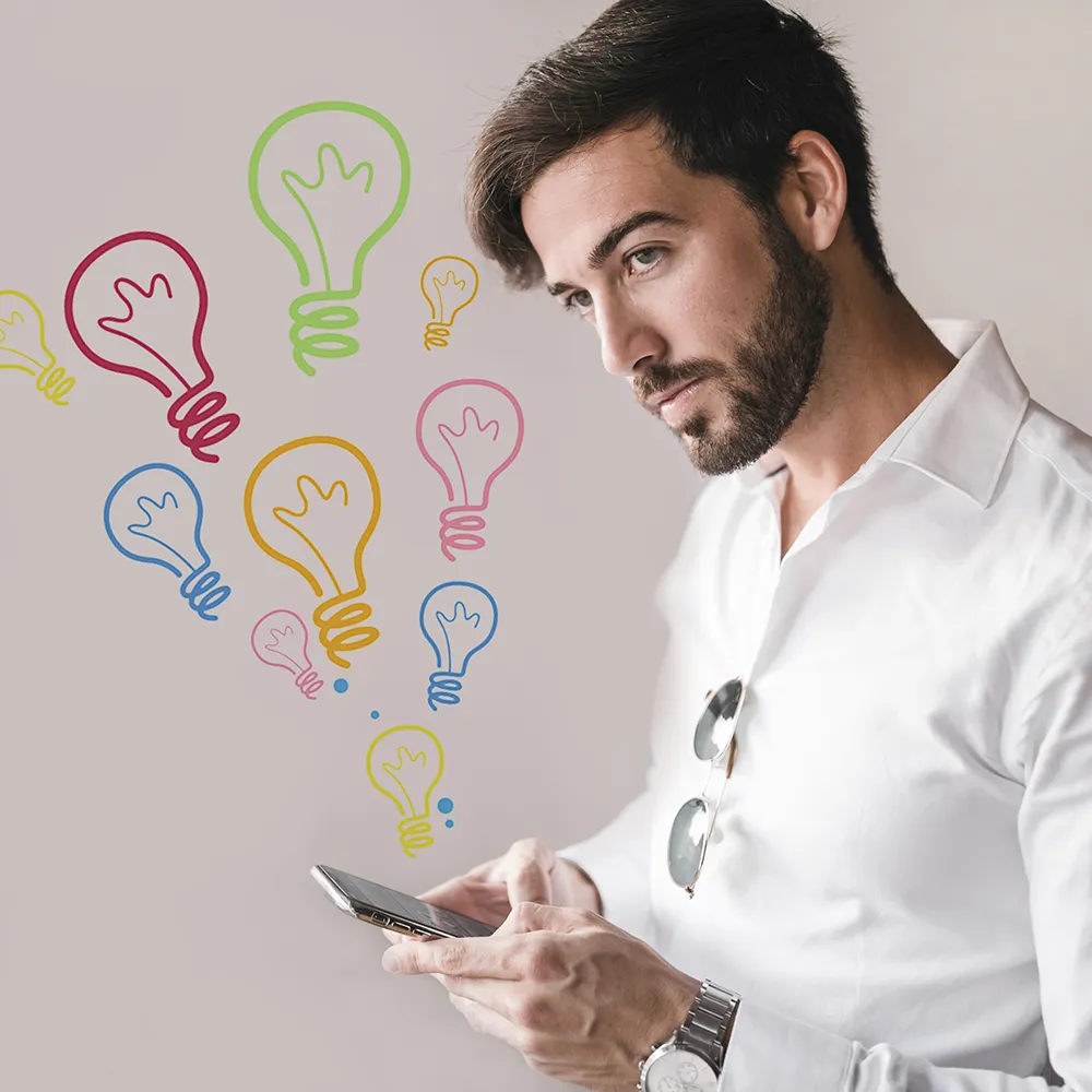 A man in a white shirt holding a smartphone, looking thoughtfully at colorful light bulb drawings floating in the air, symbolizing ideas or creativity.