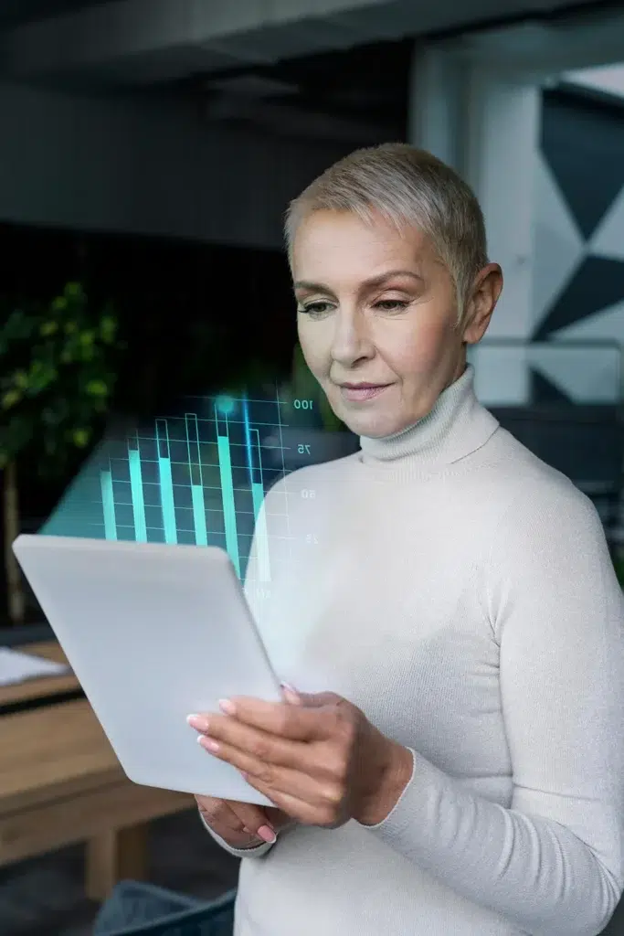 A woman with short gray hair is holding a tablet. She is wearing a light-colored turtleneck sweater and appears focused. A 3D bar graph projection is visible above the screen. The background shows a modern office setting.