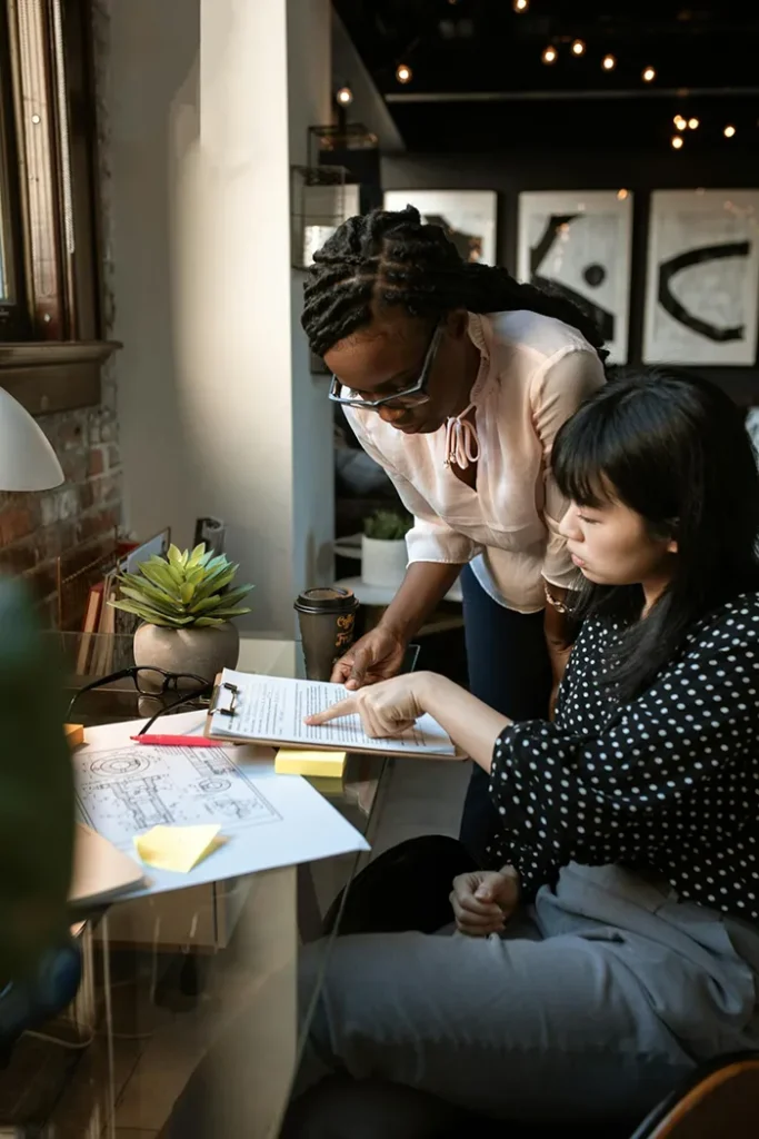 Two women work together at a desk with architectural drawings. One, standing, wears glasses and a light blouse, while the other, seated, gestures on the paper. A coffee cup and a plant are nearby in a modern office setting.