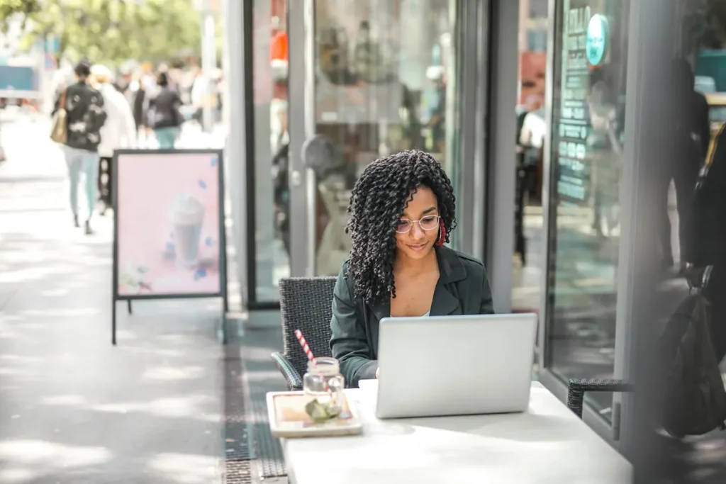 A woman with curly hair and glasses is sitting outside at a café table, working on a laptop. A beverage and a small jar of dessert are on the table. Pedestrians and a blurred street scene are visible in the background.