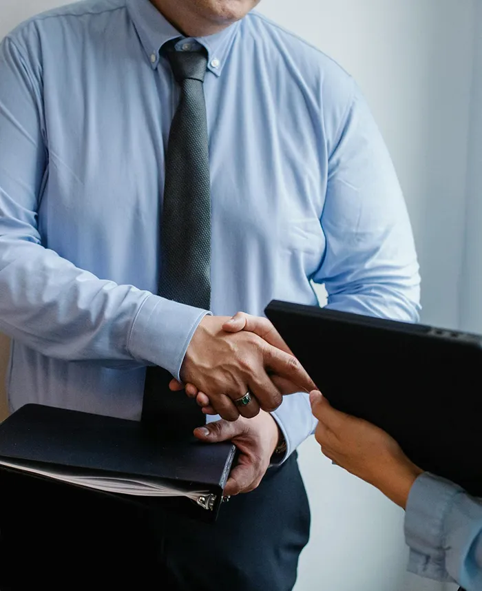 Two people in formal attire shaking hands. One is holding a black folder, and the other is holding a tablet. They appear to be engaged in a professional or business setting.