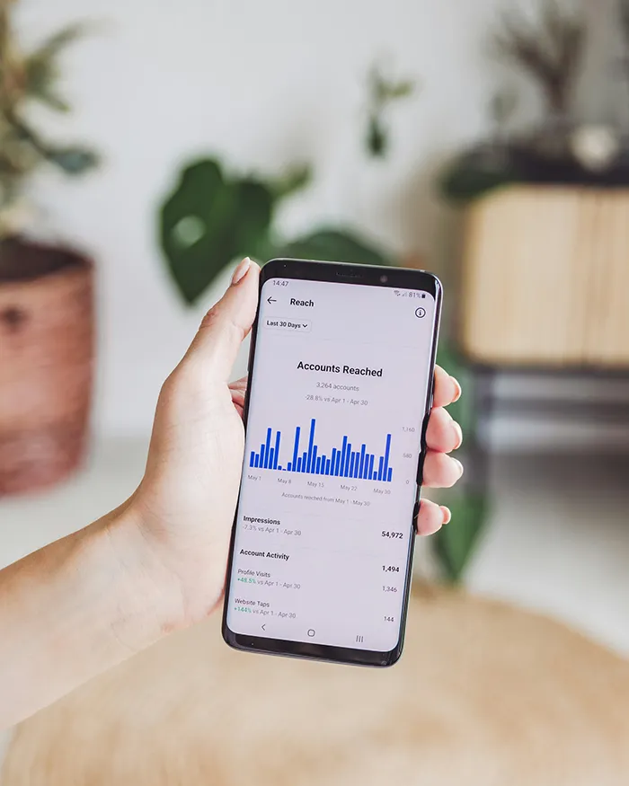 A hand holding a smartphone displaying a screen with blue bar graphs and engagement statistics. The background features blurred indoor plants and furniture.