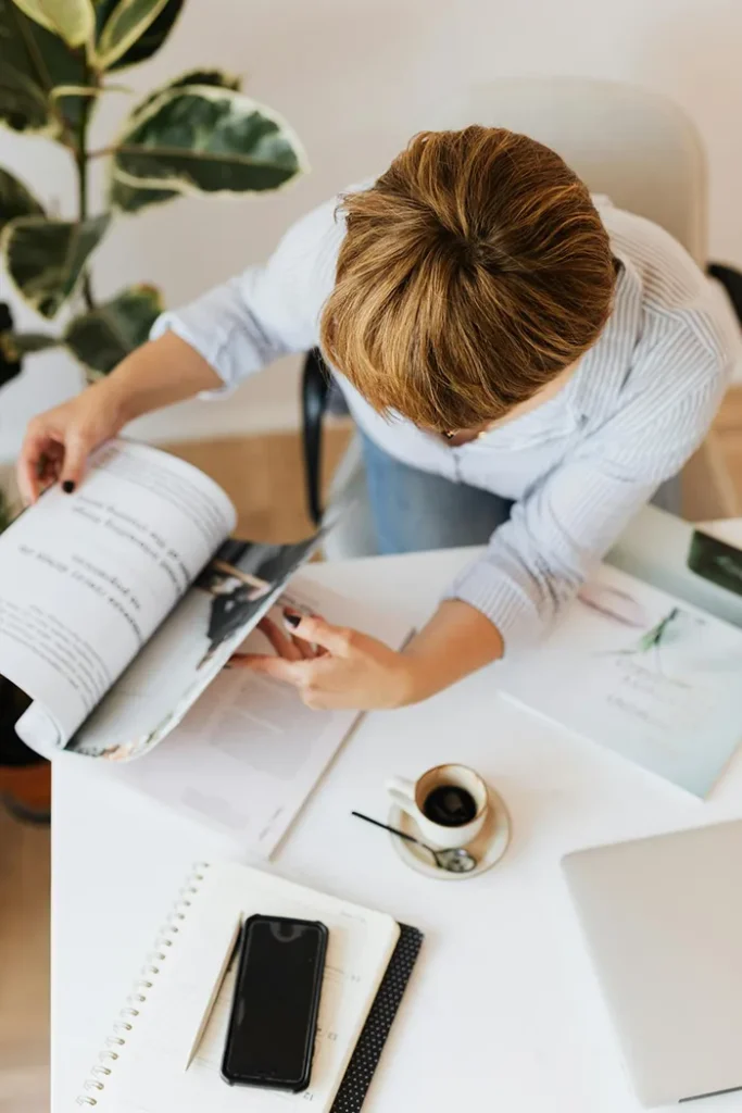 Top view of a person sitting at a desk, flipping through a magazine. The desk has a coffee cup, a smartphone, a notepad, and a laptop. A large plant is visible in the top corner, providing a touch of greenery.