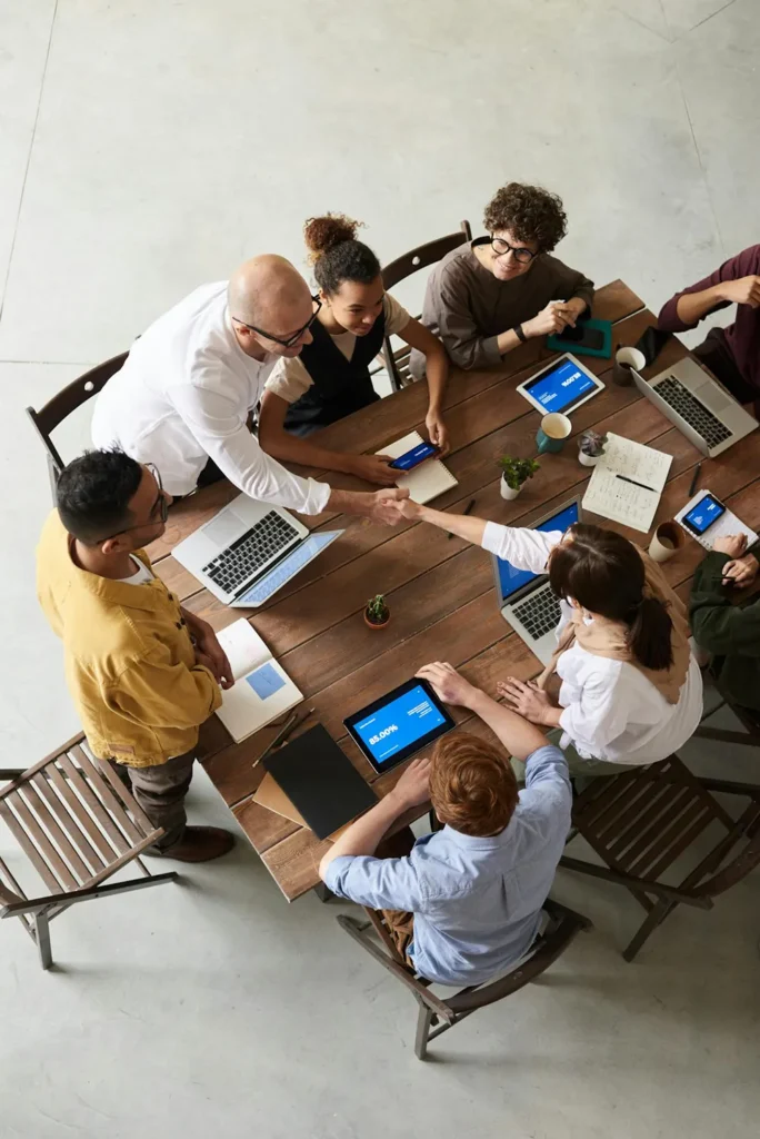 A group of people sitting around a wooden table with laptops and notebooks. Two individuals, one standing and one sitting, are shaking hands, indicating agreement or collaboration. There are small potted plants on the table.