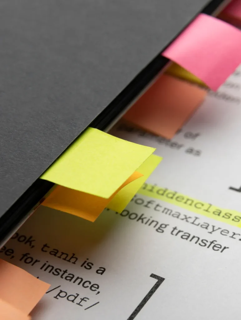 A close-up of a book with colorful sticky tabs marking pages. The tabs, in yellow, pink, and orange, protrude from the black cover. The open page beneath shows partially visible text and code elements.