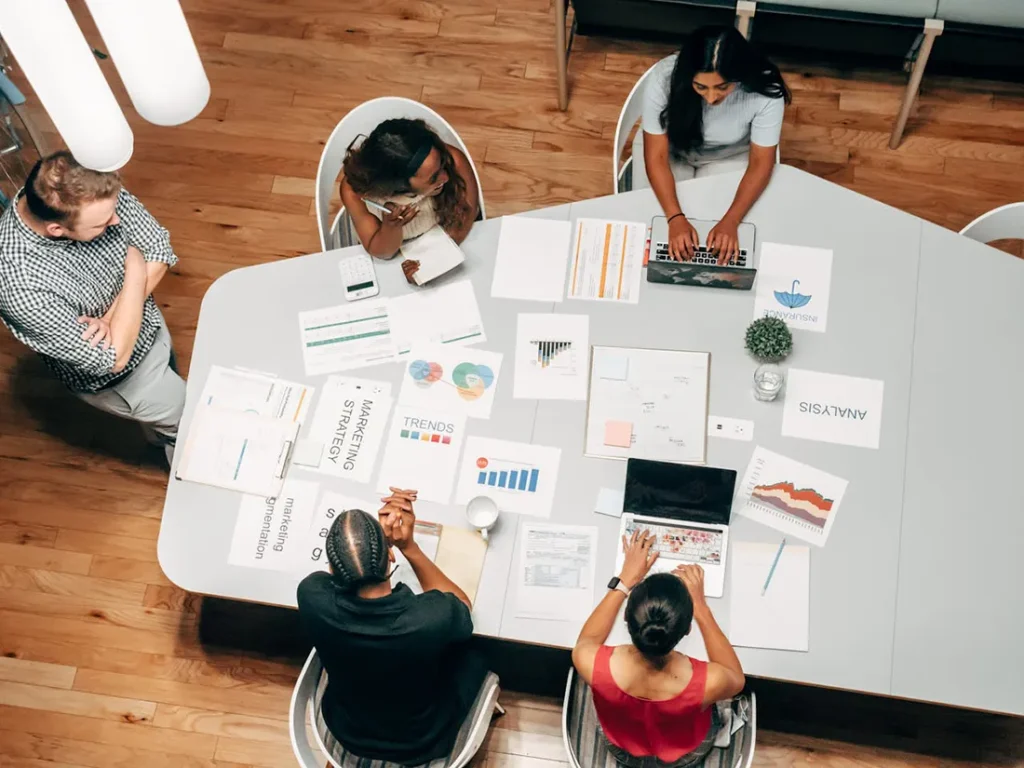 A diverse group of five people are gathered around a table reviewing documents and using a laptop. The table is covered with charts, graphs, and analytics reports. They appear to be having a meeting or brainstorming session in a modern office setting.