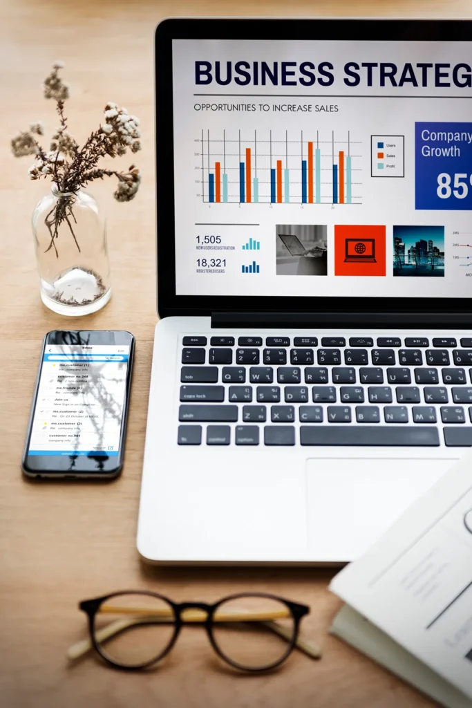 A laptop displaying a business strategy presentation with graphs and charts. A smartphone shows text and is placed beside the laptop on a wooden table. Nearby are eyeglasses, a book, and a small vase with dried flowers.