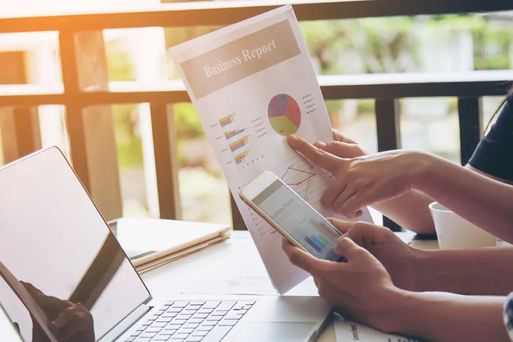 Two people at a table reviewing a business report. One points at a colorful pie chart on a printed document, while the other holds a smartphone displaying a similar chart. A laptop and a cup of coffee are visible on the table.