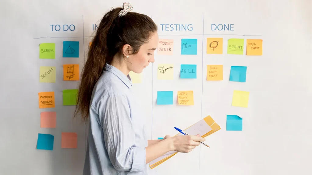 A woman with brown hair holds a clipboard and pen while looking at a kanban board. The board is divided into sections: To Do, In Progress, Testing, and Done. Various colorful sticky notes with writing are placed in each section.