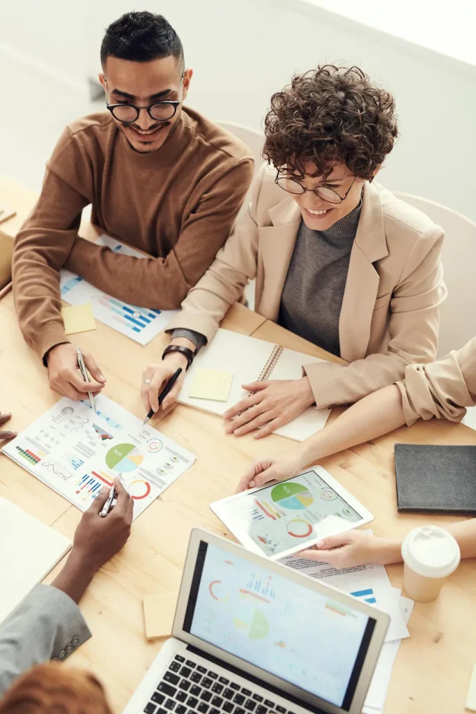 A group of professionals engaged in a meeting around a table with charts, graphs, and a laptop. Papers and a coffee cup are visible. They appear to be discussing business strategies or analyzing data.