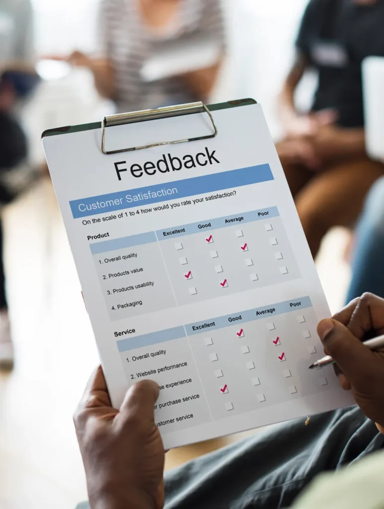 Person holding a clipboard with a customer satisfaction feedback form. The form includes categories for product and service, with checkboxes for ratings ranging from excellent to poor. Others are seated in the background, blurred out.