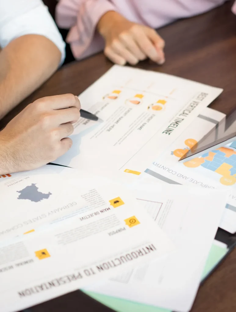 Two people are at a desk reviewing documents with charts and graphs. One person points at a colorful chart on the paper with a pen, while other documents are spread across the table.