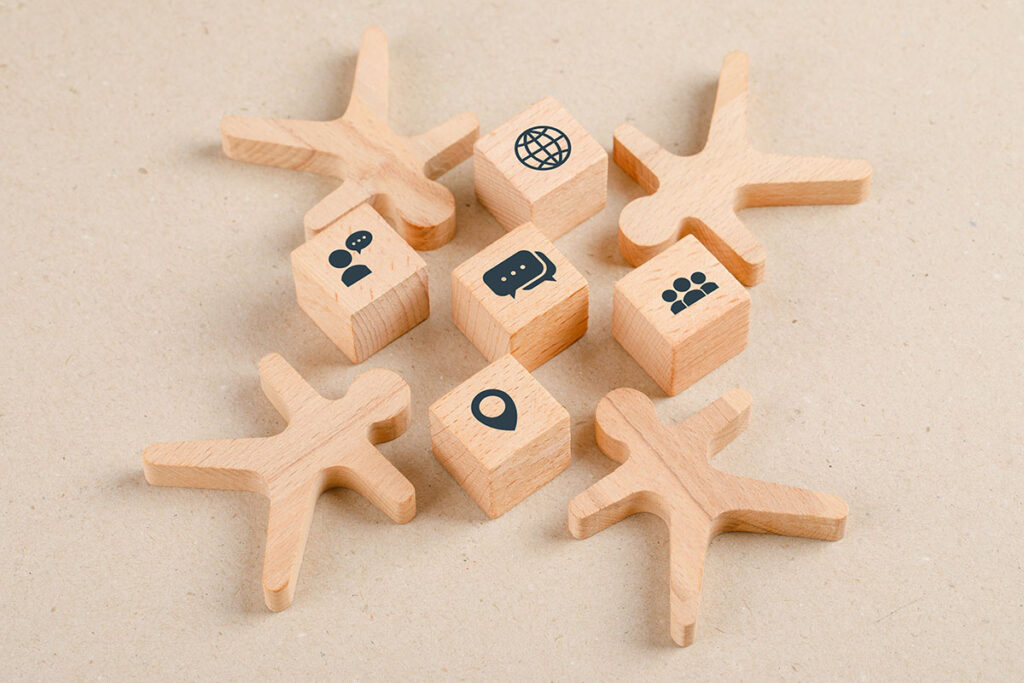 Wooden figures arranged in a circle around wooden blocks, each with icons representing communication and connection, such as a globe, chat bubbles, and people. The background is a plain beige surface.