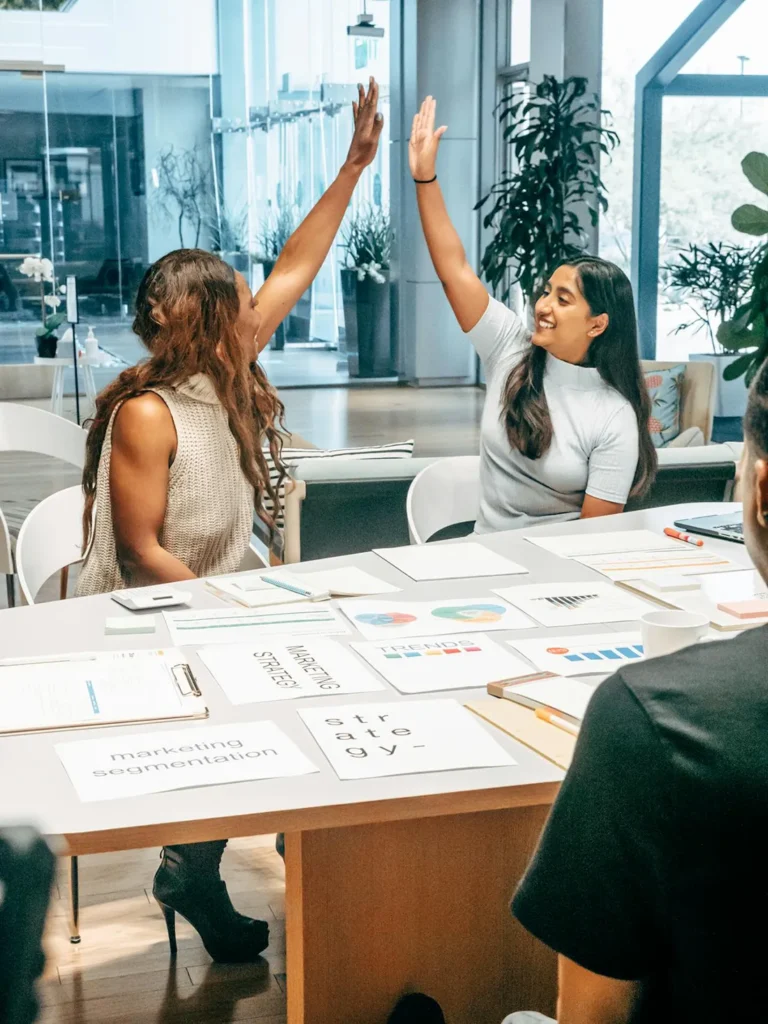 Two women seated at a conference table high-fiving each other, surrounded by papers and charts. They are in a modern office setting with large windows and plants in the background.