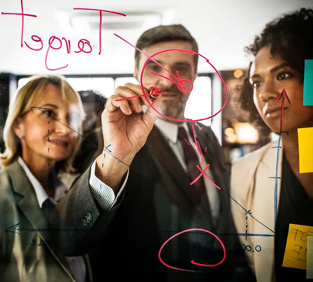 Three professionals stand behind a glass board with red and blue marker diagrams. One person is writing with a red marker. Sticky notes and the word "Target" are visible on the board. They appear to be engaged in a discussion or presentation.