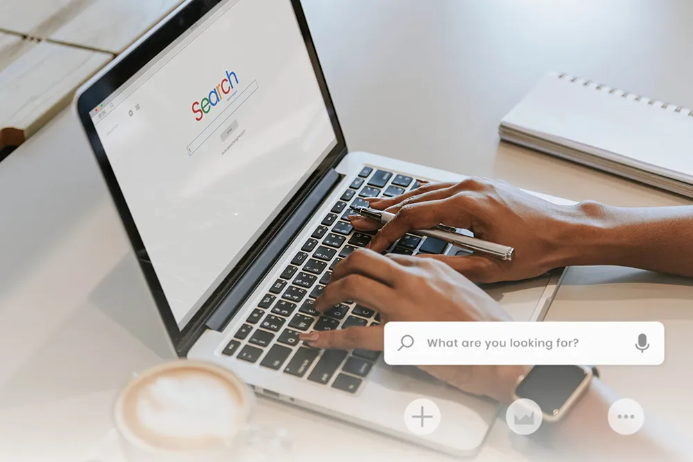 A person is typing on a laptop with a search engine open on the screen. The workspace includes a notebook, pen, smartphone, and a cup of coffee. A search box overlay with the text "What are you looking for?" is visible.