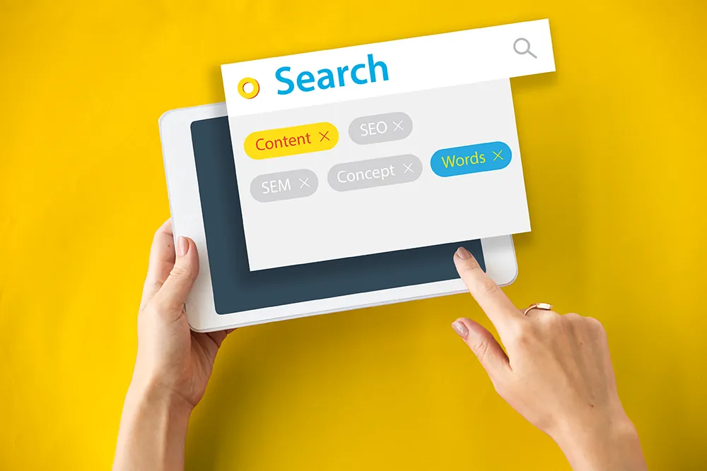 A person holds a tablet showing a search interface against a yellow background. The search window displays tagged words: Content, SEO, SEM, Concept, and Words. A finger points at the tag "Words" in the interface.