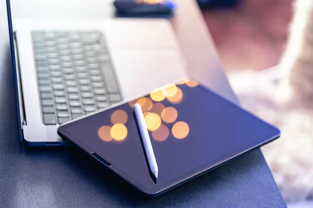 A digital drawing tablet with a stylus rests on a table next to a laptop. The screen displays abstract orange bokeh lights. The setting suggests a workspace for creative or digital art projects.