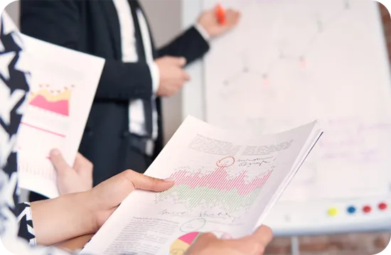 Two people in a meeting room discuss data. One holds documents with graphs and charts, while the other points to a flipchart full of diagrams. Both wear business attire, focusing on presentation materials for a SaaS SEO checklist.