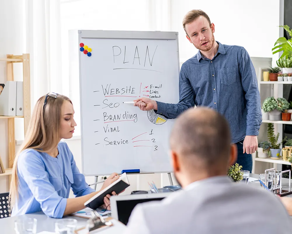 A man presents a marketing plan on a whiteboard in a bright office. Three colleagues, two men and a woman, sit around a table with laptops and notebooks. The board lists "Website, SEO, Branding, Viral, Service" under "PLAN.