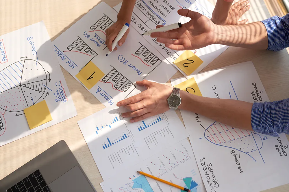 A top-down view of a collaborative workspace. Two people work with charts and graphs on paper, discussing data insights. A laptop is partially visible, along with sticky notes and a pen, suggesting a team meeting or data analysis session.