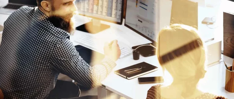 A bearded man in a checkered shirt sits at a desk with computer monitors, discussing b2b strategies with a woman whose back is turned to the camera. They are in a well-lit office of an SEO agency.
