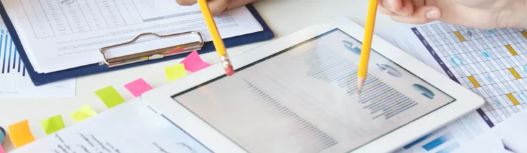 Two hands pointing with pencils at a digital tablet displaying colorful graphs and charts, possibly from a SaaS dashboard. Nearby are documents on a clipboard, additional colorful charts, and sticky notes, suggesting a business or financial meeting setting.