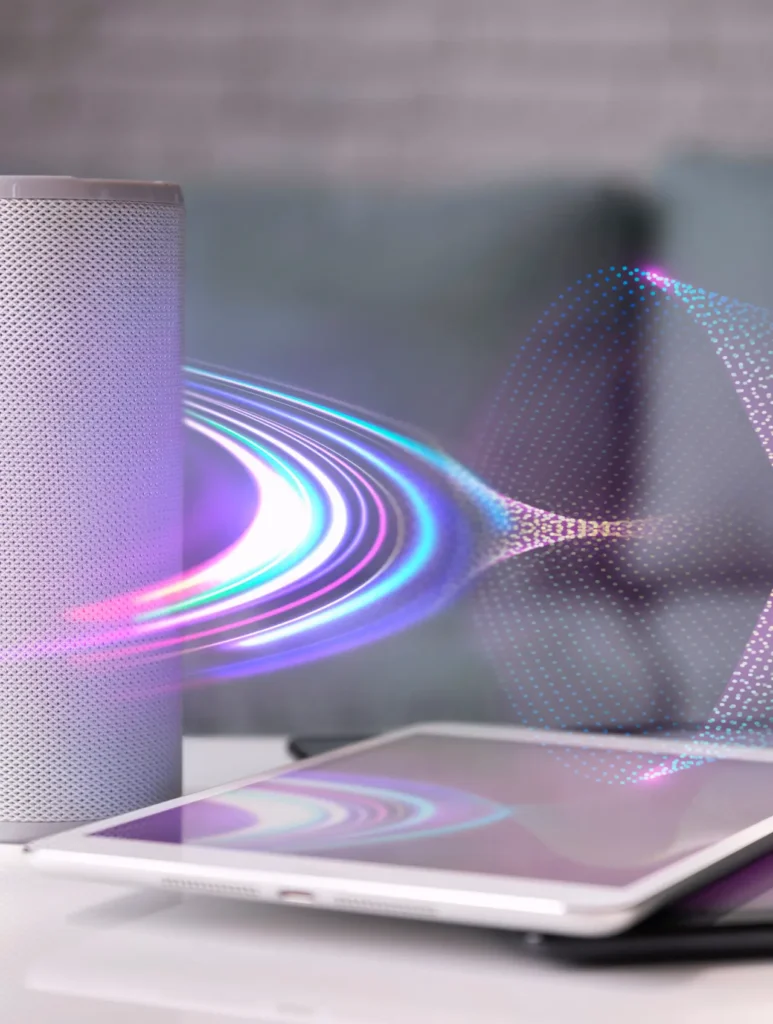 A smart speaker emits colorful, swirling sound waves towards a tablet on a table. The background is softly blurred, highlighting the interaction between the devices.