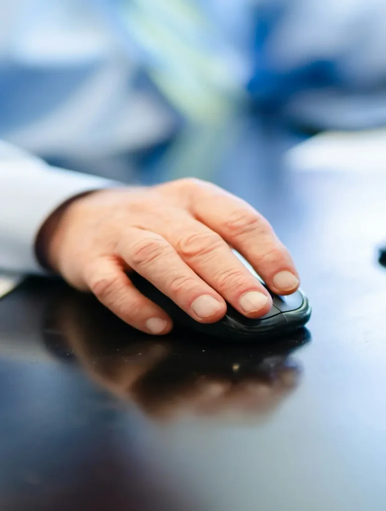 A close-up of a person's hand using a computer mouse on a dark surface. The background is blurred, suggesting a work or office environment. The person is wearing a long-sleeved shirt and a striped tie is partially visible.