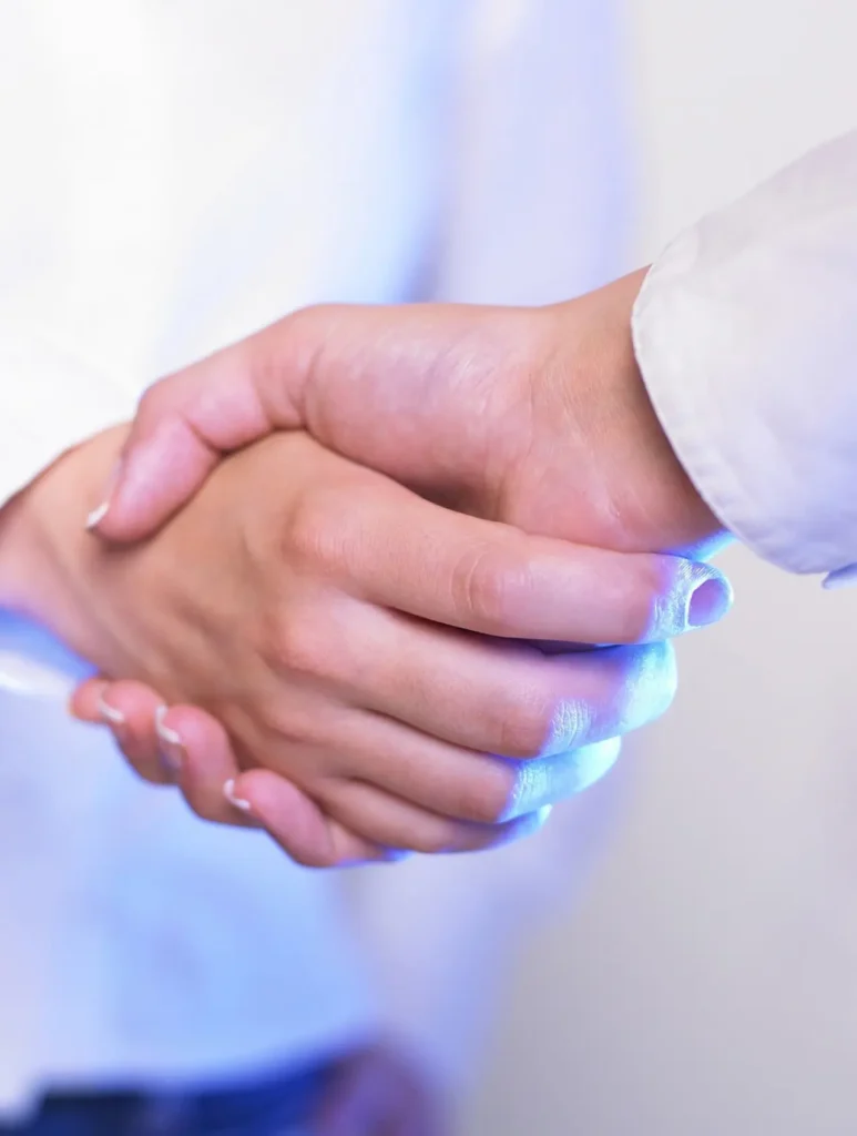 Two people in white shirts shaking hands, symbolizing agreement or partnership. The image focuses on their hands against a softly lit, neutral background.