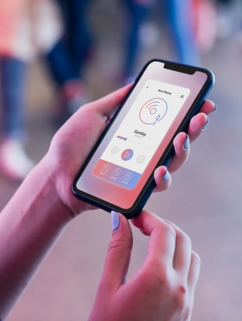 A person holding a smartphone displays a weather app with a red and blue design. The screen shows the temperature, a weather icon, and the day "Sunday." The person's nails are painted blue. The background is blurred.