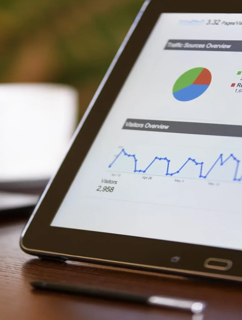 A tablet displaying analytics, featuring a colorful pie chart, a line graph, and the text "Visitors: 2,958" on the screen. The device is on a wooden surface next to a pen, with a blurred background.