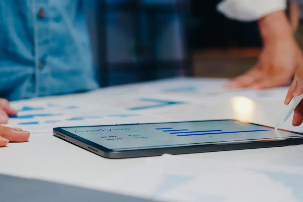 A digital tablet displaying bar charts and graphs is placed on a table. A person is using a stylus to interact with the screen, while another person observes. Papers with charts are spread across the table.