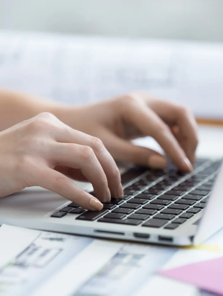 Close-up of hands typing on a laptop keyboard, with papers and plans spread out on the table in the background.
