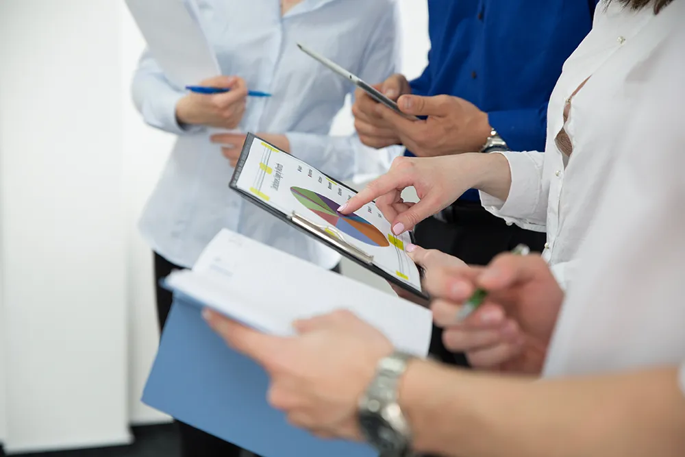 A group of four people standing together, holding documents and clipboards, discussing a chart on one clipboard. The image captures a professional setting focused on collaboration and data analysis.
