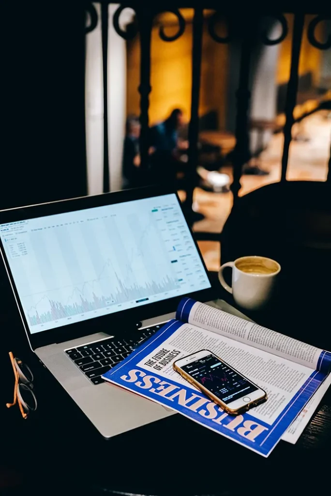 A laptop displaying a stock chart is on a table alongside a business magazine, a smartphone, a cup of coffee, and a pair of glasses. The setting appears to be indoors with a blurred background.