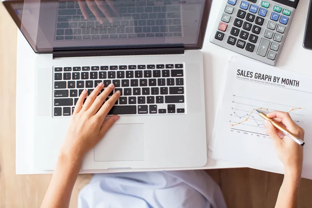 A person uses a laptop with their right hand writing on a "Sales Graph by Month." Calculators and papers are on the desk, indicating work or study involving data analysis or financial tasks.