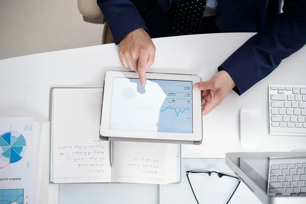 A person in a suit points at a tablet screen displaying graphs and charts, indicating data analysis. The workspace includes an open notebook with handwritten notes, a keyboard, and glasses.