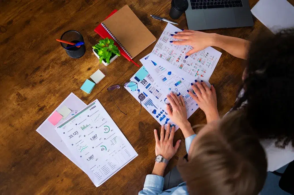 Top view of a wooden table with two people reviewing charts and graphs on paper. A laptop, notebook, sticky notes, a small plant, and a pair of glasses are also on the table.