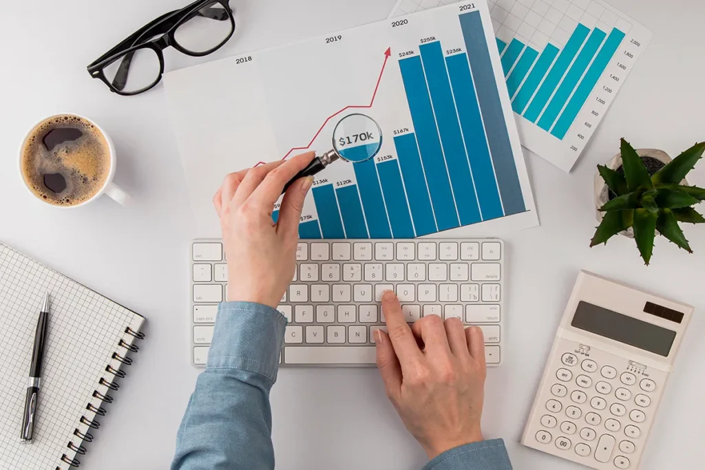 A person wearing a blue shirt uses a keyboard, holding a magnifying glass over a bar graph with a rising trend line and "$170k" label. Nearby are a coffee cup, glasses, notepad, calculator, and a small plant.
