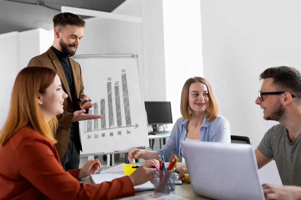A group of four people in a modern office setting engage in a discussion around a table. A standing person gestures towards a whiteboard displaying a bar graph, while others sit with a laptop and notebooks, actively listening and smiling.