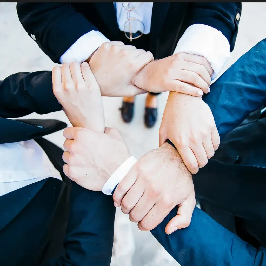 Four people in business attire standing in a circle, each holding the wrist of the person next to them, symbolizing teamwork and unity.