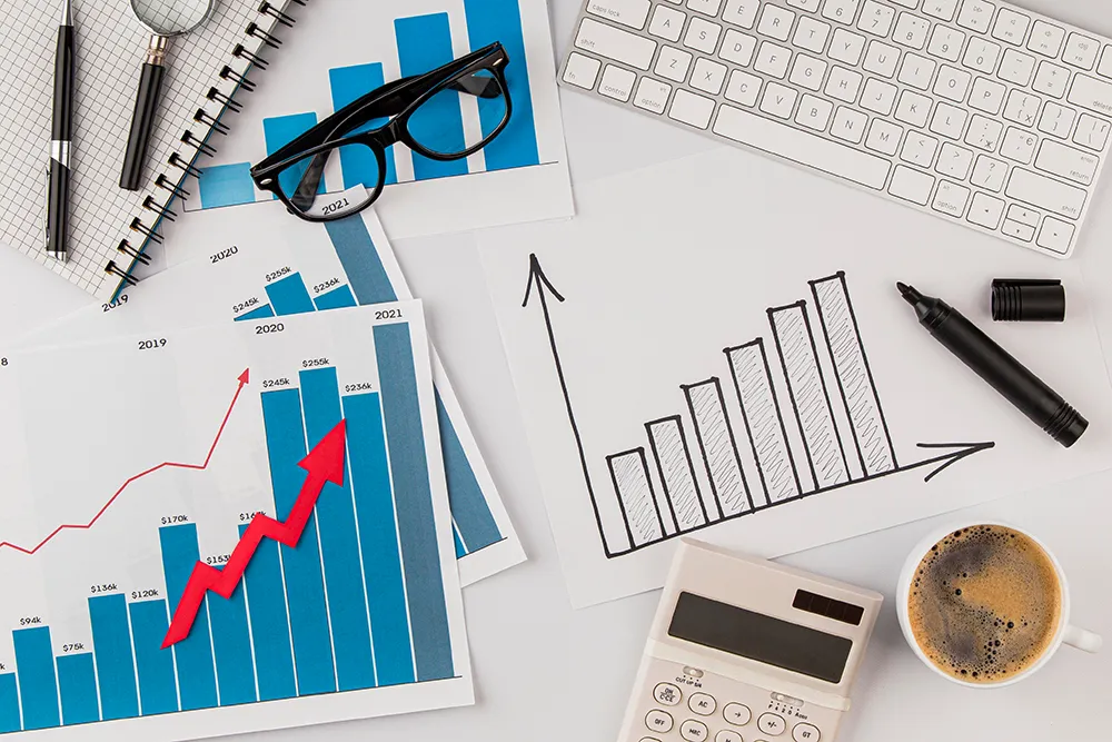 A desk with bar and line graphs, a calculator, a cup of coffee, glasses, a keyboard, and a magnifying glass. Charts indicate rising trends, and a black marker is next to a hand-drawn graph on paper.