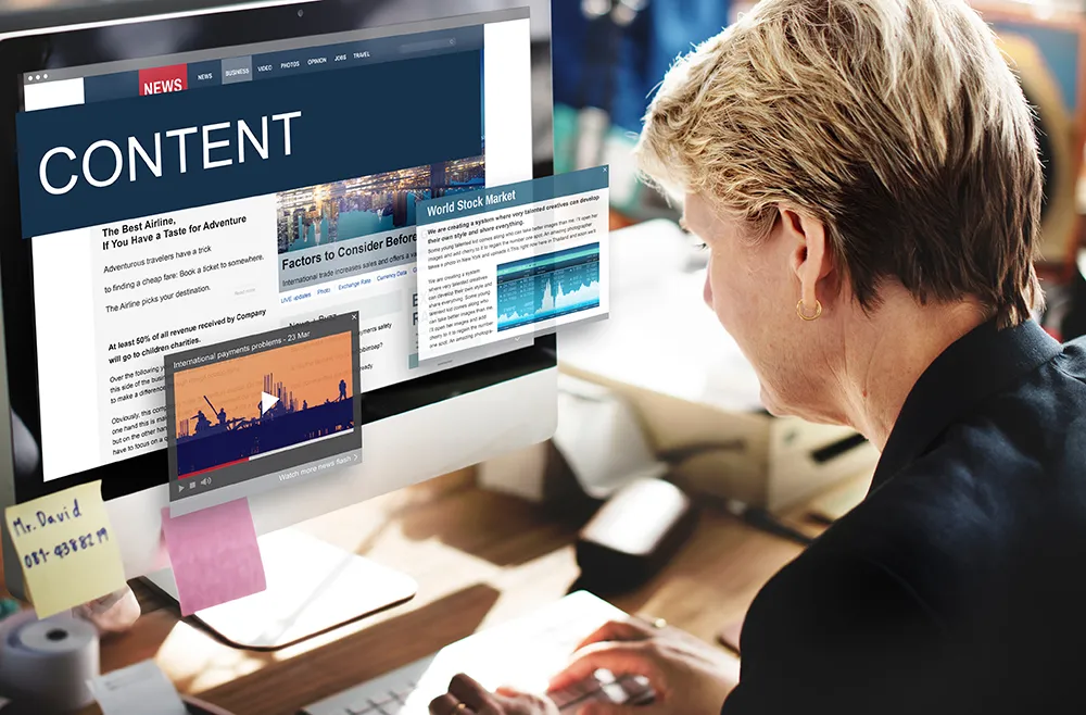 Person at a desk using a computer, viewing various digital content including news articles, a video, and stock market data. The screen displays large text "CONTENT" with a menu on the side. Papers and office supplies are scattered on the desk.